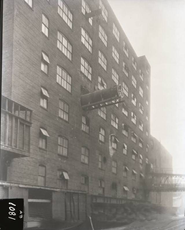 A large piece of equipment is being manoeuvred out of building onto railway car below, 1924