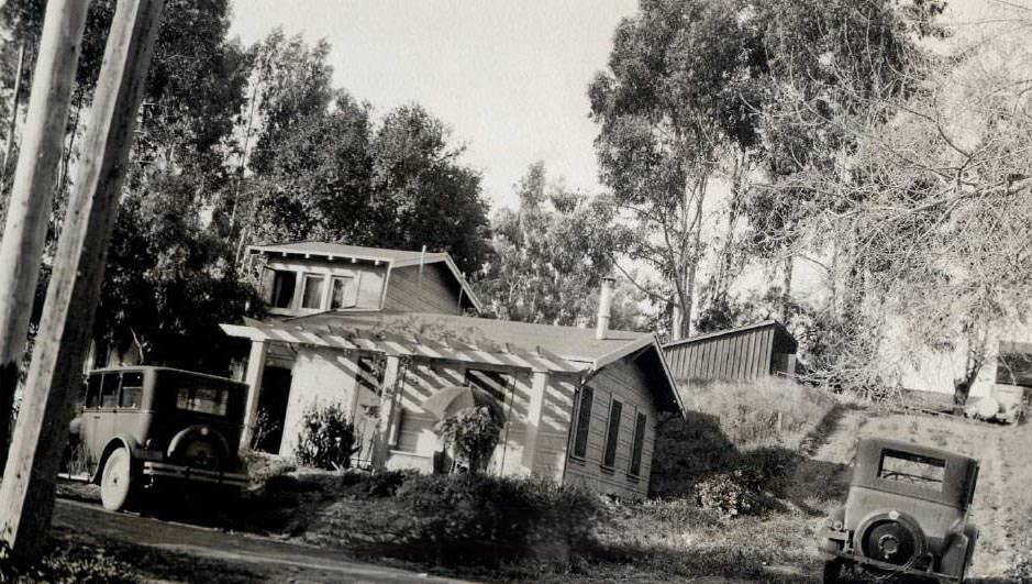 Small house nestled up against wooded hillside. Two cars are parked alongside the dirt roads, 1920s