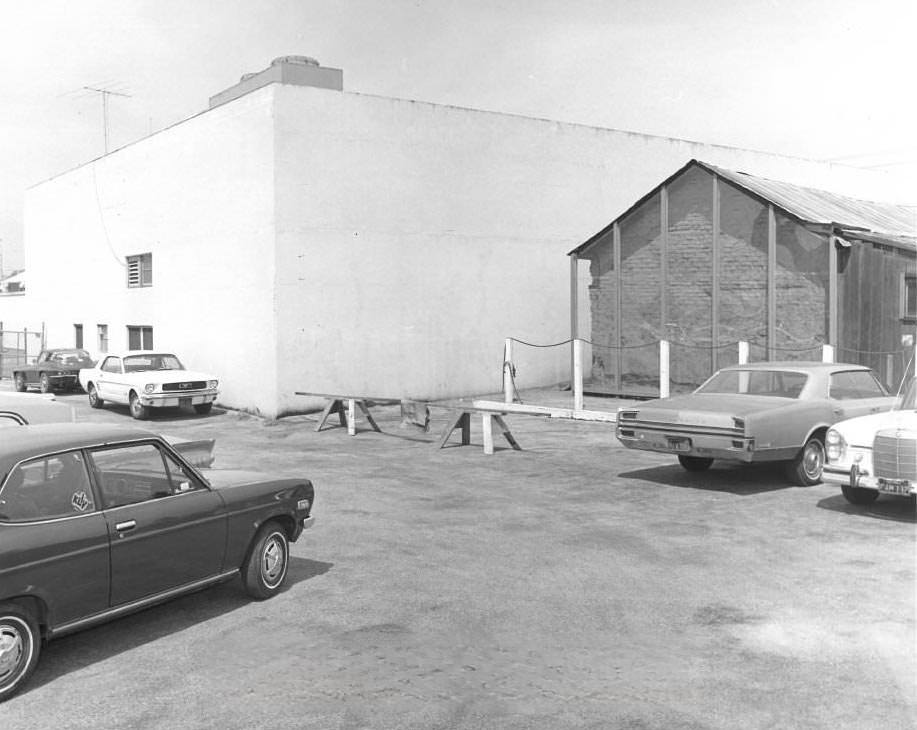 Cars parked in a lot next to the side of the Peralta Adobe, with the Lusardi building behind it, 1976