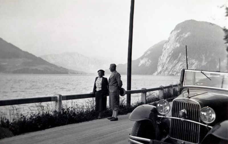 A middle-aged couple posing with a Steyr XX Cabriolet on a gravel road running along the bank of a lake on a bleak summer's day, 1930