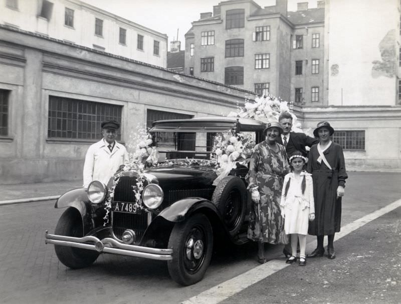 Four members of a middle-class family and a chauffeur posing with an abundantly decorated Steyr 30 Landaulet Typ 45 in an urban backstreet, 1932