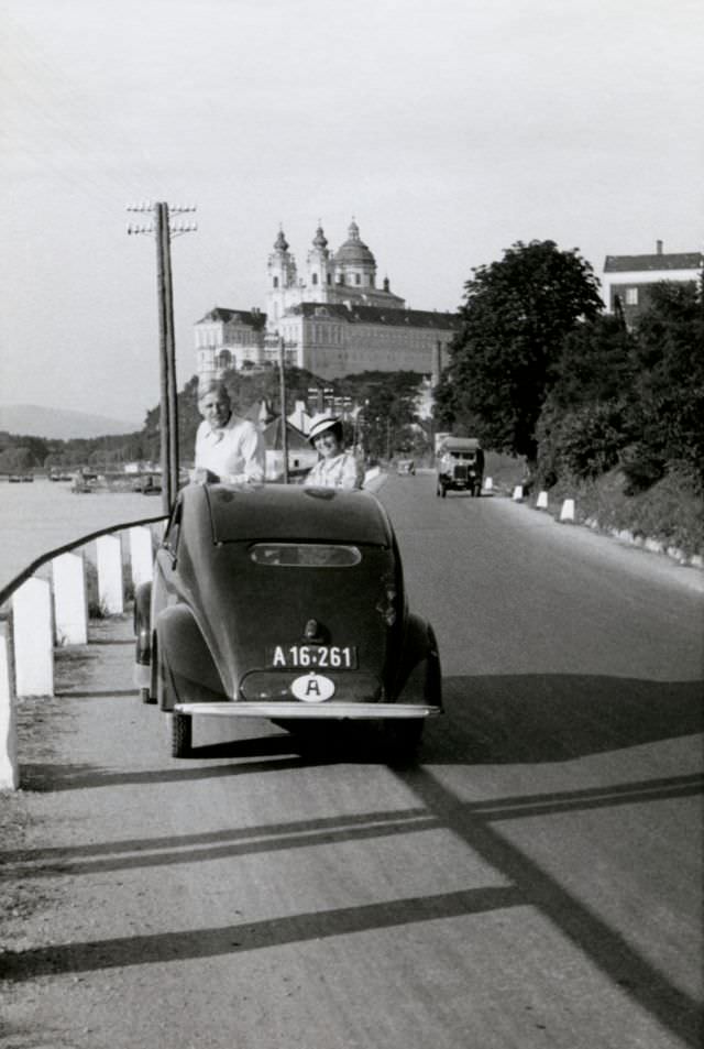 A couple posing with a Steyr 50 on the side of the road in the town of Melk on the bank of the Danube with the famous monastery visible in the background, 1937