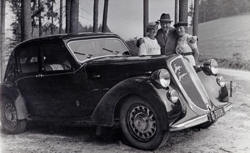 Two ladies in white dresses and a fellow wearing a suit and a hat posing with a Steyr 125 Super in the countryside, 1937