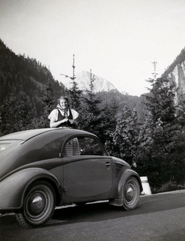 A cheerful young lady peeking out of the sunroof of a Steyr 50 on a mountain road, 1938