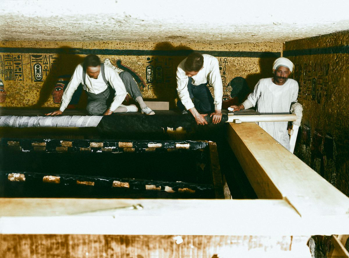 Howard Carter (left), Arthur Mace and an Egyptian workman standing on scaffolding, roll back the linen pall (Carter no. 209). Tutankhamun's Tomb, 30th December 1923