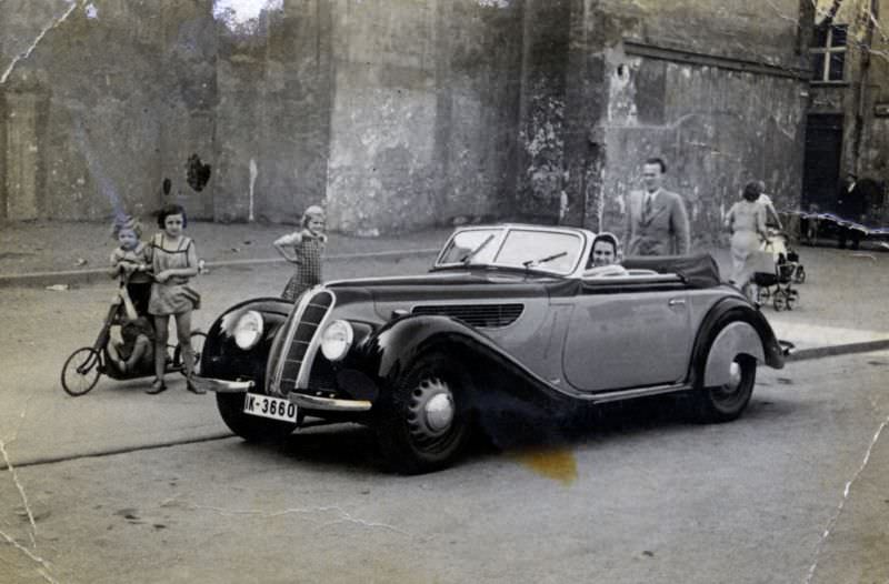 A cheerful lady posing in the driver's seat of a BMW 327 Cabriolet in a rundown urban backstreet, 1938.