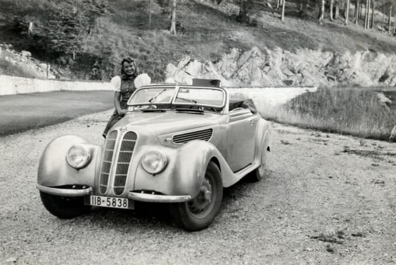 A cheerful lady posing with a BMW 327/28 Cabriolet convertible on a mountain road in summertime, 1938.