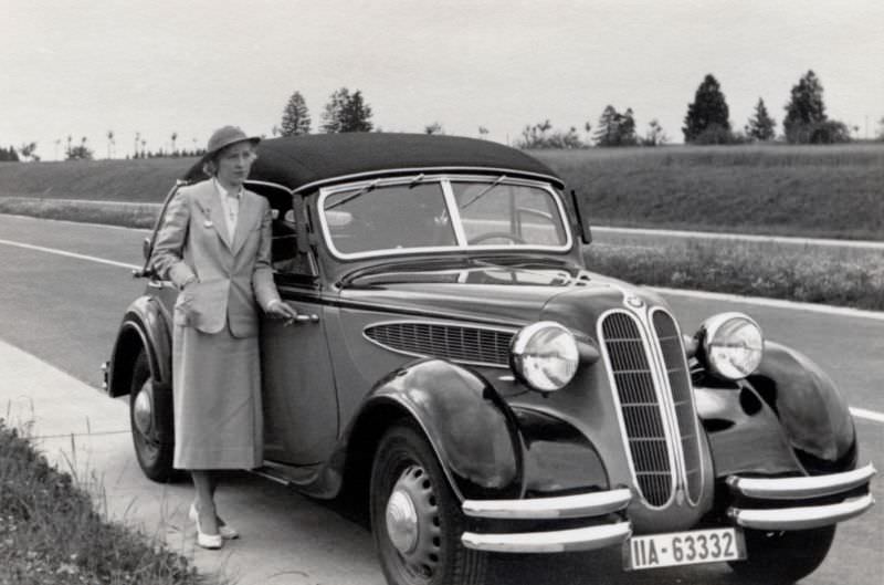 An elegant lady in a female suit posing with a BMW 326 Cabriolet on a stretch of newly-built Autobahn, 1938.