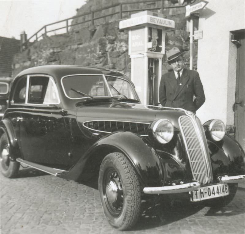 A fellow wearing a double-breasted pinstripe suit and hat posing with a BMW 320 Limousine 2 Türen next to a 'Bevaulin' petrol pump, 1939.