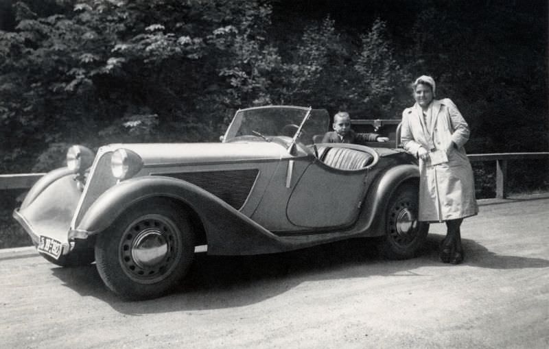 A lady wearing a light-colored coat and a flying helmet posing with a BMW 315/1 on a gravel road in the countryside, 1948.