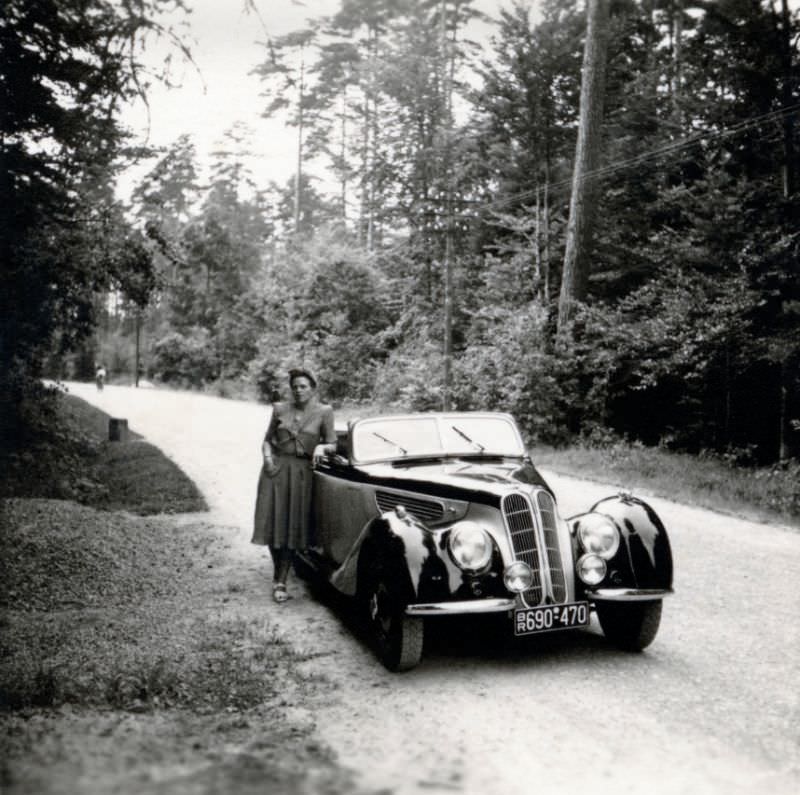 A young lady dressed in the fashion of the forties posing with a BMW 327/28 Cabriolet convertible on a gravel road in the countryside, 1949