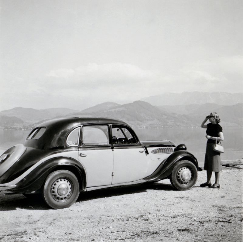 A blonde lady posing next to a two-tone BMW 326 by the side of an Alpine lake in summertime, 1950