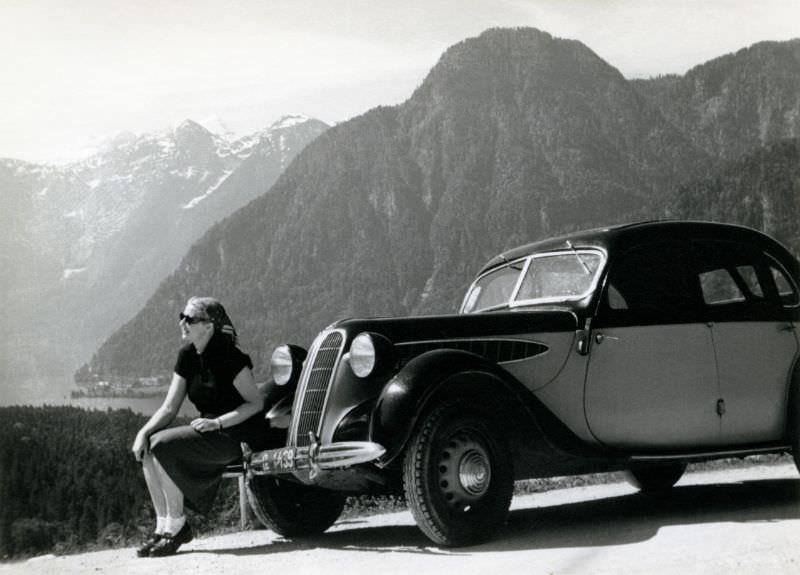 A blonde lady posing on the bumper of a pre-war BMW 326 on an Alpine road overlooking a lake, 1950