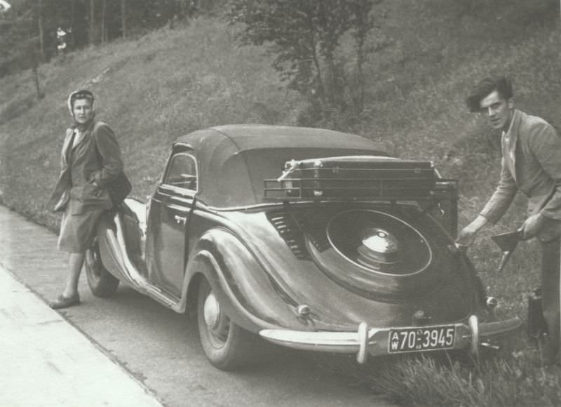 A fellow with a wild haircut refueling his BMW 327 Cabriolet, while his girlfriend or wife is looking on, 1950.