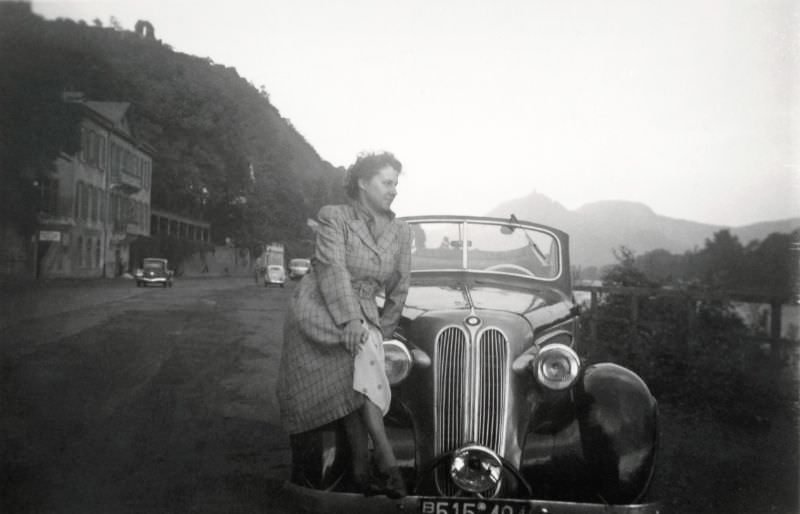 A young lady in a checkered coat posing on the fender a BMW 320 Cabriolet on the side of the road on a bleak summer's day, 1950.