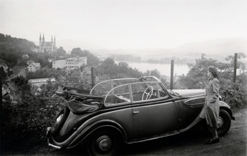 A young lady in a checkered coat posing with a BMW 320 Cabriolet convertible on a hillside overlooking the Rhine Valley, 1950.
