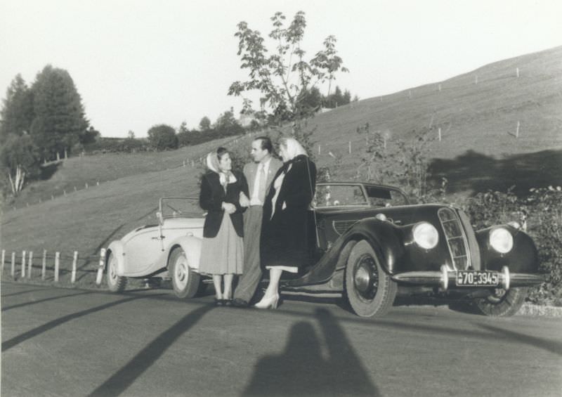 Two ladies and a gent posing with two convertibles on a mountain road in late-afternoon sunshine, 1950.