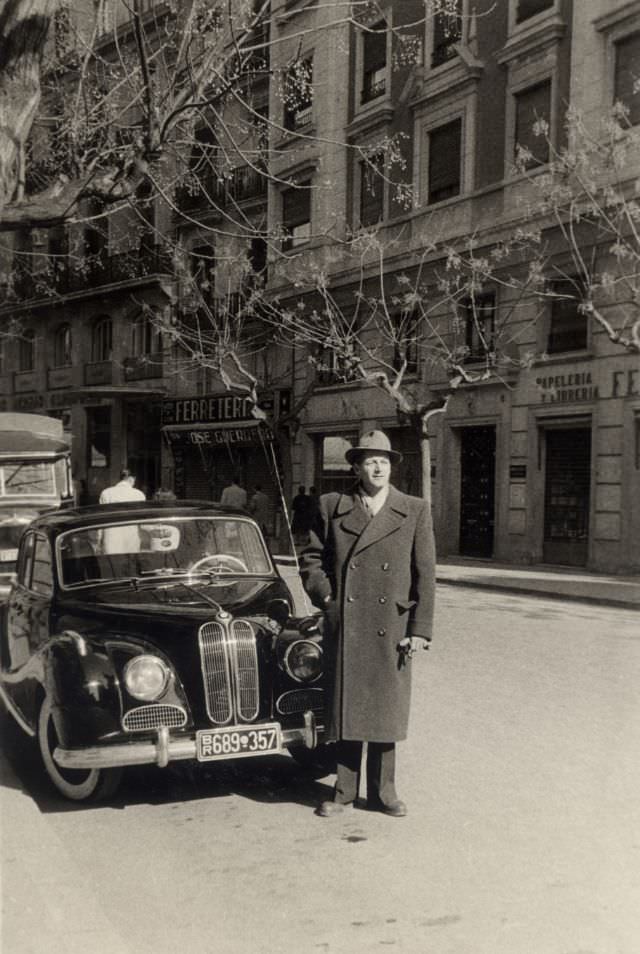 A fellow in a double-breasted coat posing with a BMW 501 on a sunny winter's day, 1954.