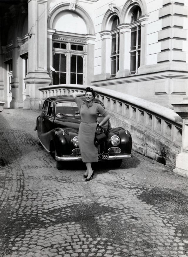 A brunette lady in a 3/4-length dress posing with a BMW 501 on the cobbled driveway of a mansion or palace,  1955