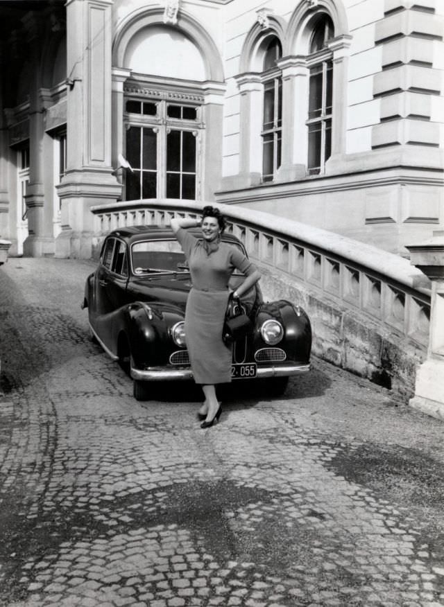 A brunette lady in a 3/4-length dress posing with a BMW 501 on the cobbled driveway of a mansion or palace, 1955