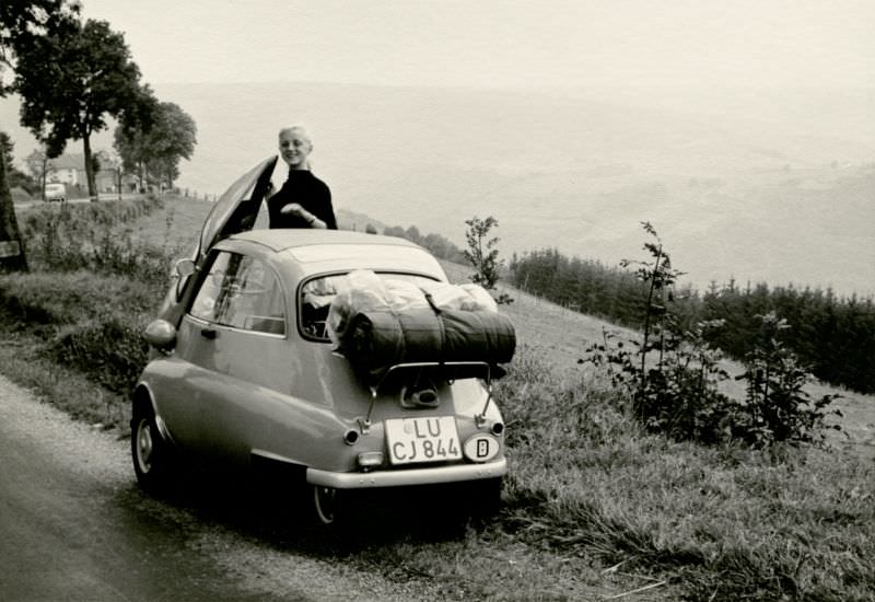 A young lady posing with a BMW Isetta on the side of a tree-lined country road, 1958.