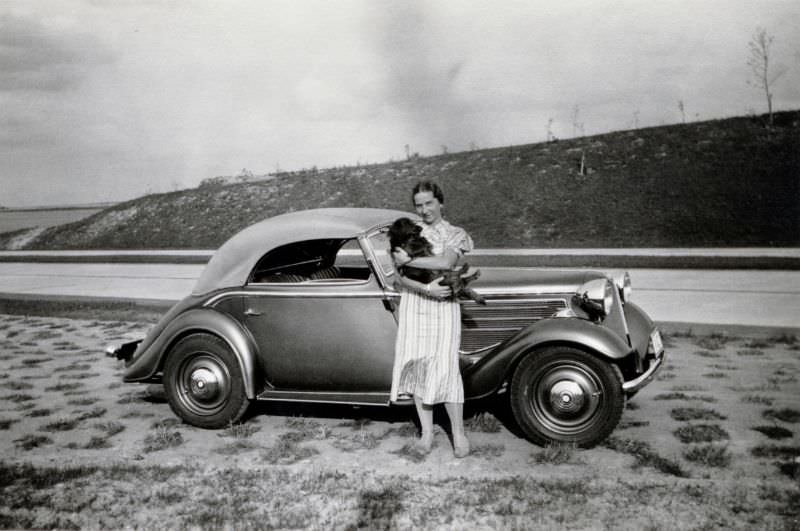 A lady in a striped summer dress posing with a BMW 319 Sport-Cabriolet convertible on a stretch of newly-built Autobahn, September 5, 1936