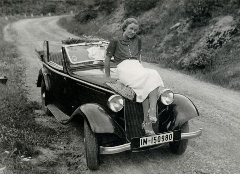 A young lady dressed in the fashion of the 1930s posing with a BMW 303 Cabriolet on a gravel road in the countryside, 1936.