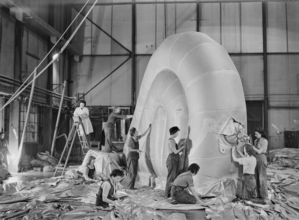Female workers, including a number wearing dungaree overalls, construct and assemble the fin of a barrage balloon in a factory hanger in England during World War II on 2nd December 1942.