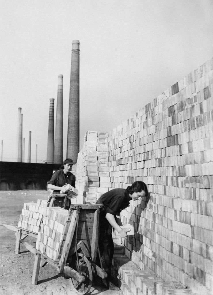 Smoke stack chimneys of the London Brick Company form the backdrop to two women war workers stacking new bricks for the construction industry and the war effort on 1st September 1942 at Stewartby in Bedfordshire, United Kingdom.