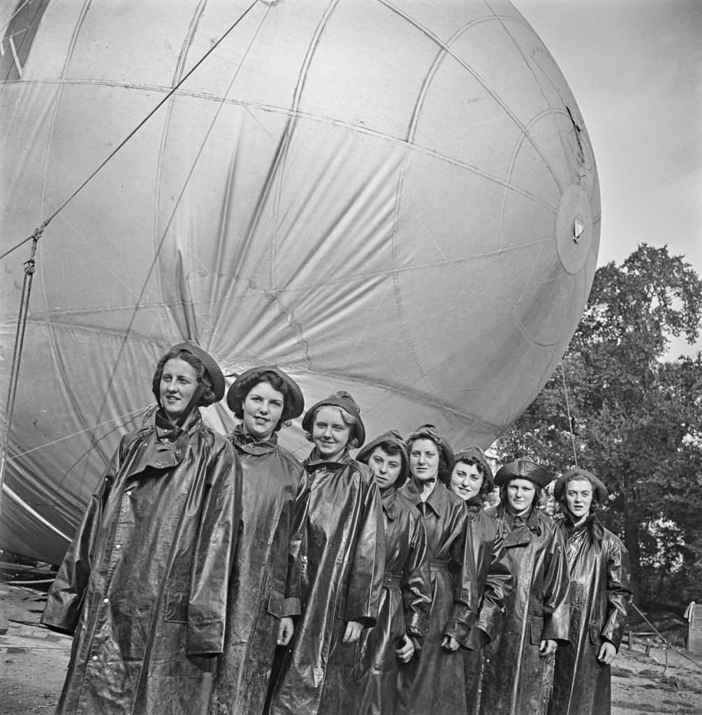Members of the Women's Royal Air Force (WRAF) from RAF Balloon Command stand together in their wet weather waterproof oilskins in front of their allocated barrage balloon tethered in Grosvenor Gardens, London. 11th September 1941.