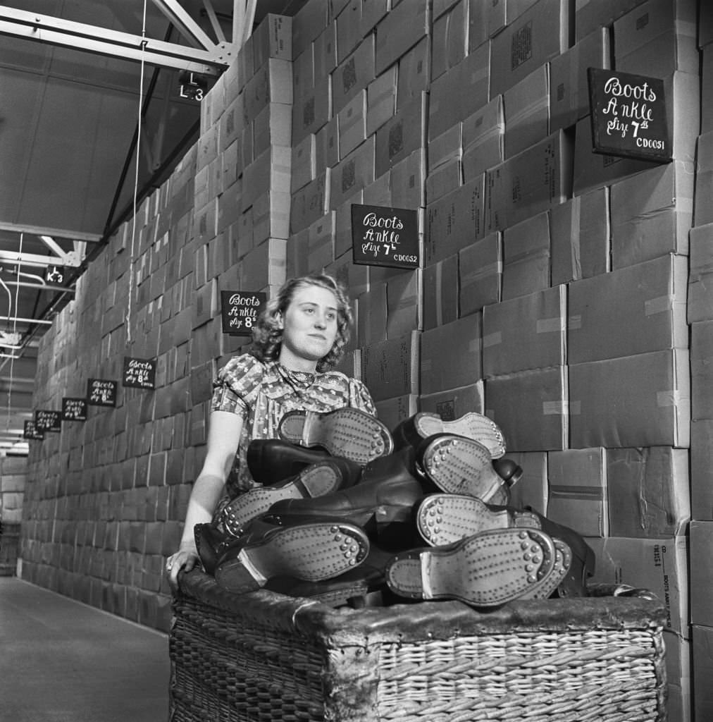 A female warehouse worker pushes a wicker basket full of Britsh Army standard issue Ammunition boots past stacks of hobnail boot boxes at a Central Ordnance Depot for British Army uniform and equipment in England during World War II.