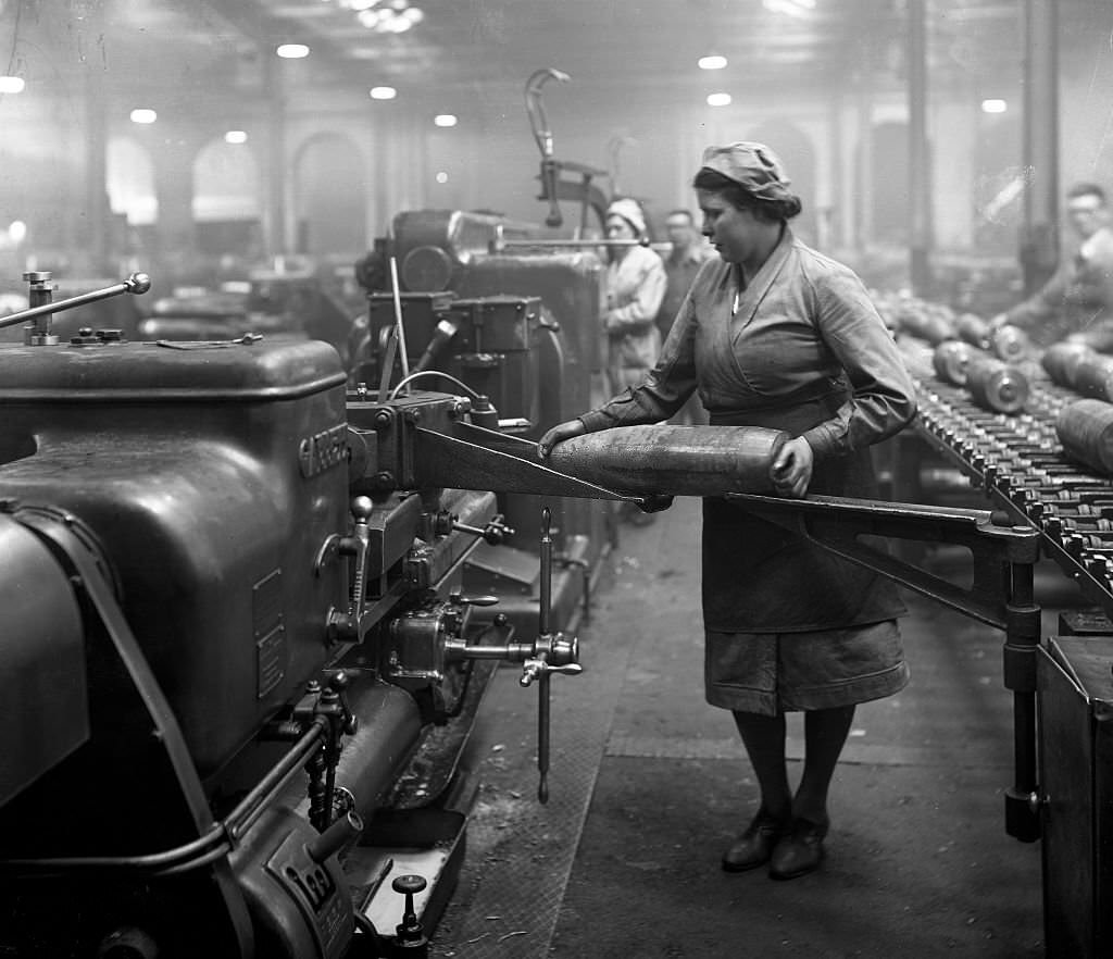 Works photograph of female employee transferring a 6 inch shell from the bench to a bore fuse hole machine, Female employee transferring a 6 inch shell from the bench to a bore fuse hole machine.