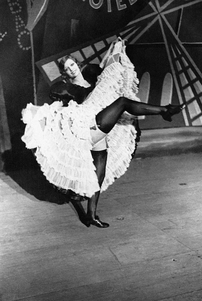 A Cancan dancer takes a break by raising her leg on stage in front of the wings of a windmill, 1900