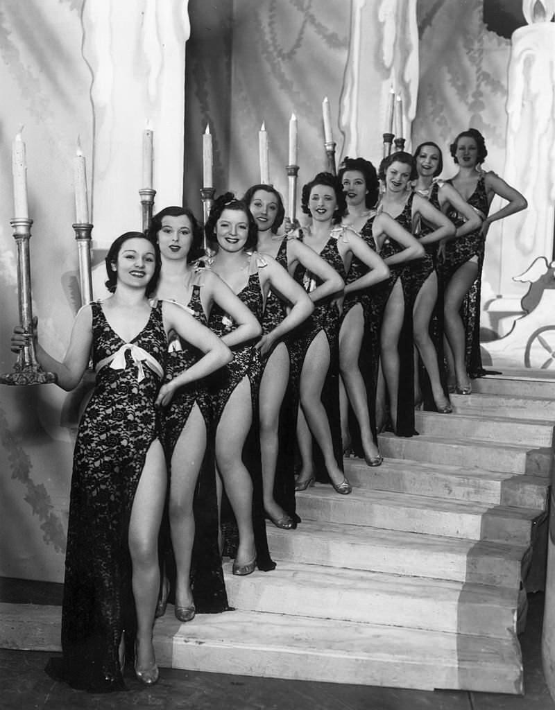 Holding candles in large candlesticks chorus girls from the 'Folies de Can-Can' pose on a staircase, 1938