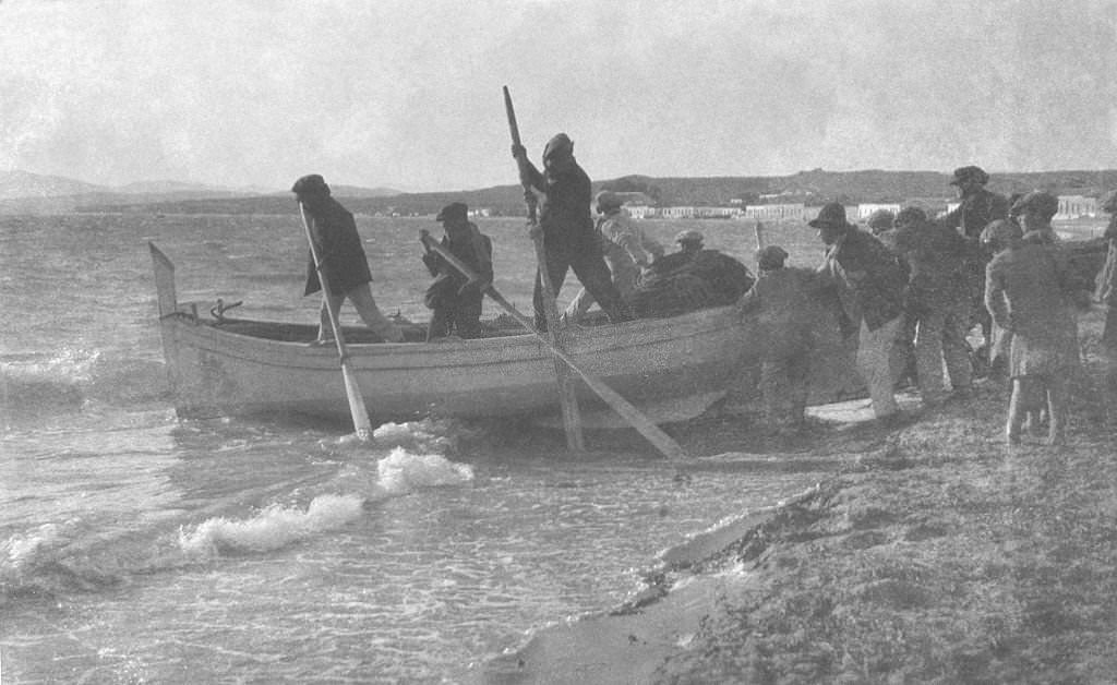 Group of men in a fishing boat holding oars as their boat is pushed into the sea from the shoreline by another group of men as children watch on, Egypt, 1910.