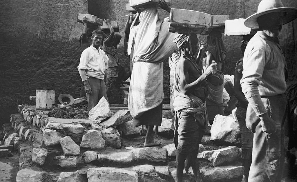 An Archaeologist stands among rocks and stones of an archaeological dig as a group of tribal workers carry boxes on their heads from the site Egypt, 1910.
