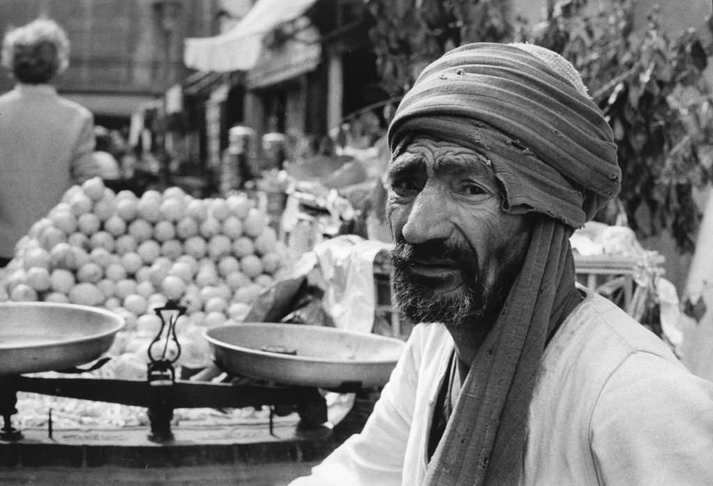 A fruit wither in front of its scales at a stand in the Khan El Khalili bazaar in Cairo, 1972