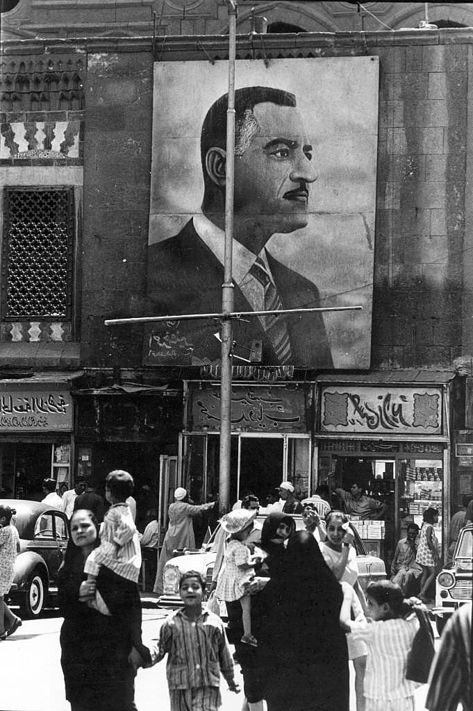 Under a large banner that depicts Egyptian President Gamal Abdel Nasser, pedestrians, some with children, walk on a city street, Cairo, Egypt, 1970.