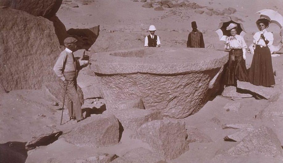 Several men and women stand around a large stone sarcophagus, still in the rock quarry near Aswan, where it was left unfinished thousands of years ago, 1900