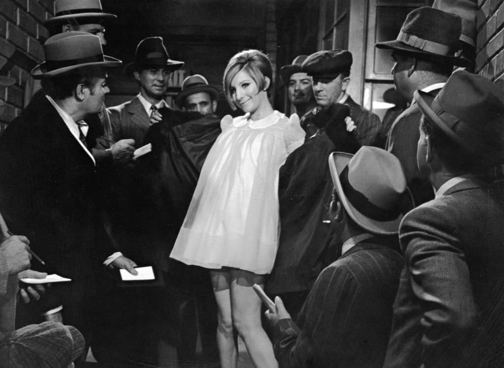 Barbra Streisand opens her coat to reveal a yellow nightgown as reporters gather around her in a still from the film 'Funny Girl' 1968