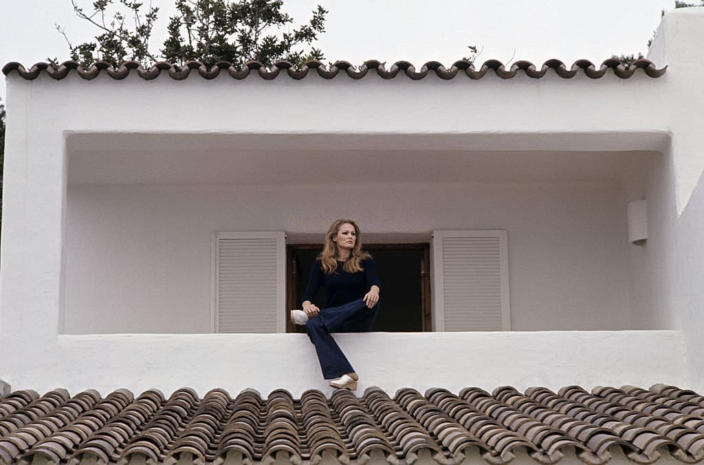 Actress Ursula Andress, in jeans and a dark sweater, sitting on the low wall of her villa's balcony, Ibiza, 1970s