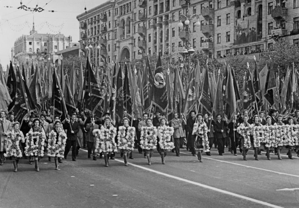 Ukrainian women wearing floral costumes, which spell out an unspecified phrase, as flagbearers, one with a flag of the Communist Party of the Soviet Union with an image of Lenin,