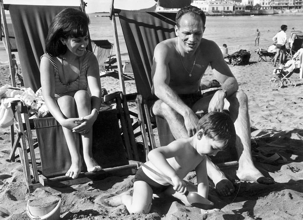 British boxing champion, Henry Cooper sitting on a beach at Las Palmas de Gran Canaria, Gran Canaria with his wife and son, 1965