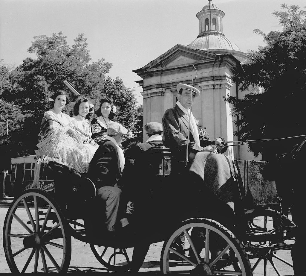 Several women ride in a horse-drawn carriage next to the Hermitage of San Antonio de la Florida, in Madrid during the 1960s.