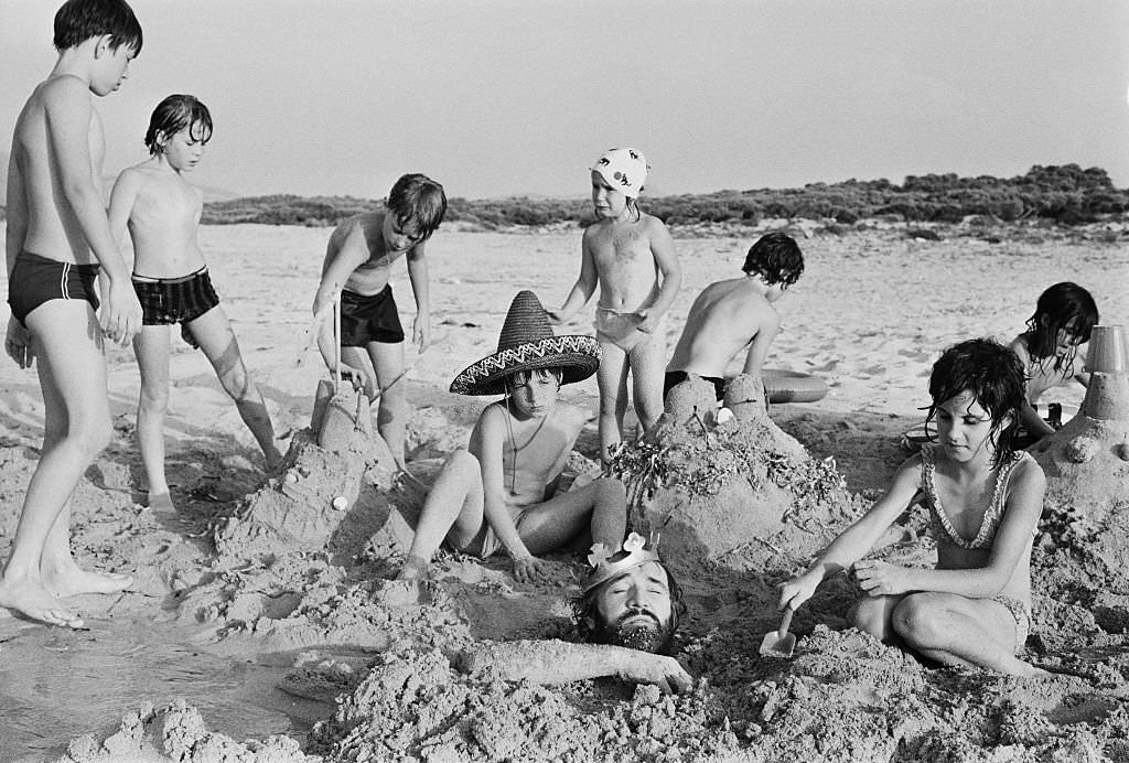 Irish actor Richard Harrisbeing buried in sand by a group of children, whilst on holiday in Majorca, 18th August 1971