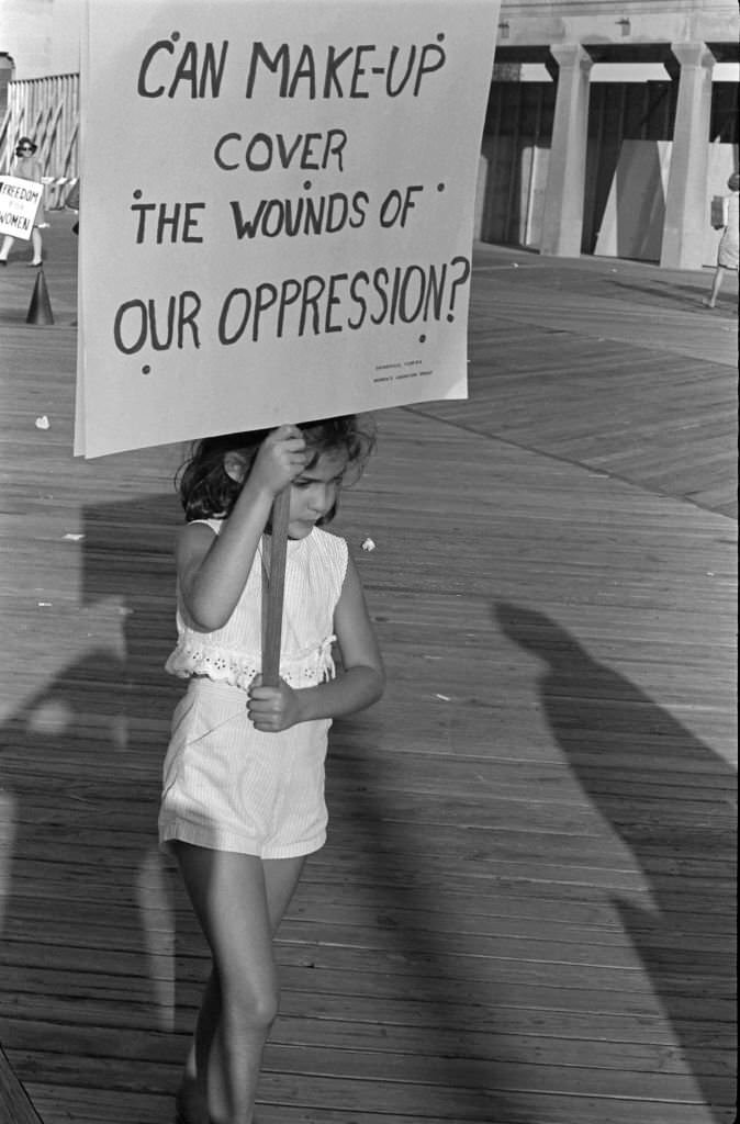A young demonstrator carries a poster that reads 'Can Make-Up Cover the Wounds of Our Oppression' as, with others, she protests the Miss America beauty pageant, Atlantic City, 1968.