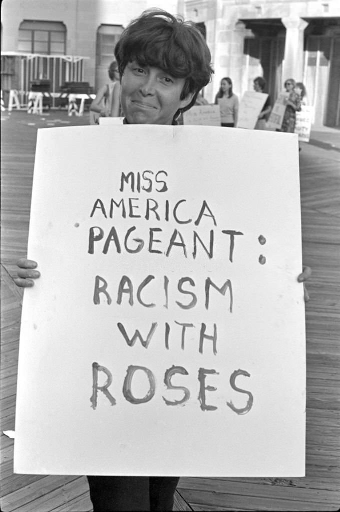 A demonstrator as she holds a poster that reads 'Miss America Pageant: Racism with Roses' as she poses on the Atlantic City Boardwalk during a protest against the Miss America beauty pageant, Atlantic City, 1968.