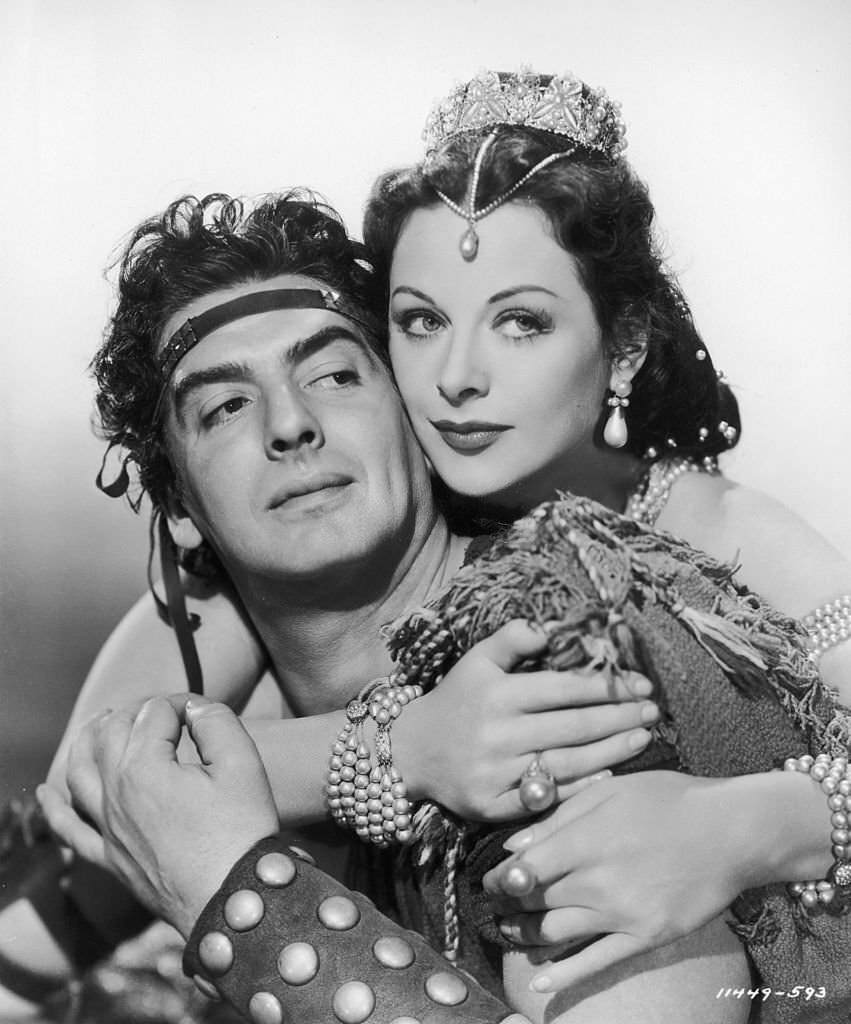Hedy Lamarr wearing a costume with pearls, leans against Victor Mature wrapping her arms around his shoulders from behind.