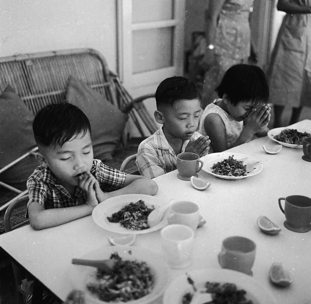Three children in Singapore in prayer before a meal at a Red Cross school for disabled children, 1964