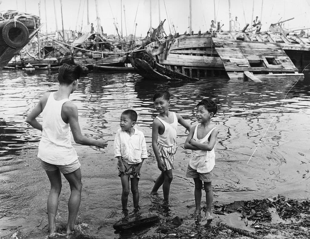Singaporean children play in the harbour, while behind them there is little activity on the boats, 1963